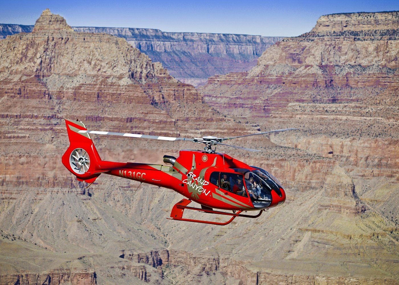 Red helicopter flying over the Grand Canyon with passengers visible inside, showcasing the stunning canyon landscape.